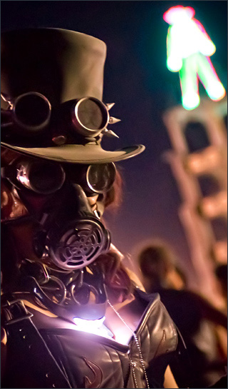 Woman in a top hat, spiked goggles, gas mask, glowing necklace, chain, and leather top, in front of a bright humanoid shape that stands atop a tall tower.