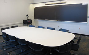 Large, wide chalkboard in a lecture room with a rectangular table and 16 moveable chairs placed around it. 