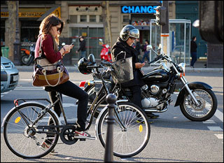 A woman on a bicycle and a man on a motorcycle are both looking down at phones in their hands while stopped in traffic.