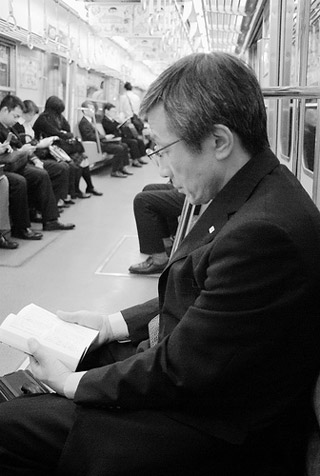 A man in a black coat sits on a subway and reads a book.