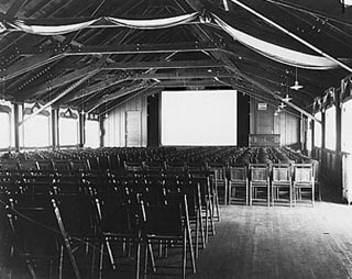 A black and white photo of movie hall with rows of wodden chairs.