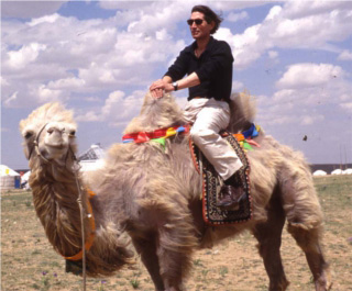 Photo of Dr. Wheatley riding a camel in front of a row of white tents.