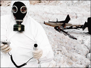A photograph of a person wearing a hazmat suit and holding a radiation detector, and standing in front of a banner landscape with a destroyed object in the background.
